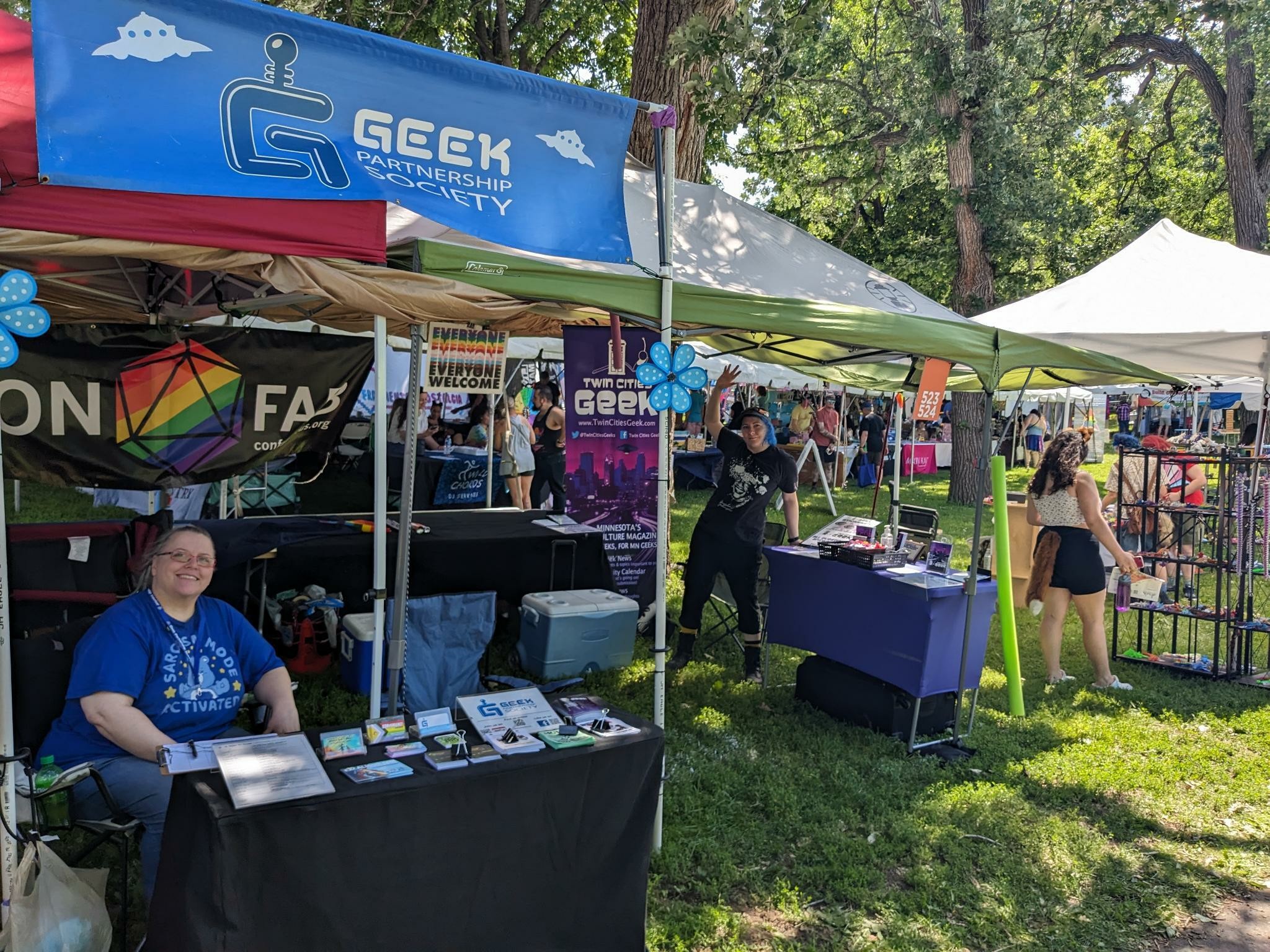Two people at the Geek Partnership Society booth outside on a sunny day, featuring a GPS banner in the foreground and a Twin Cities Geek banner in the background
